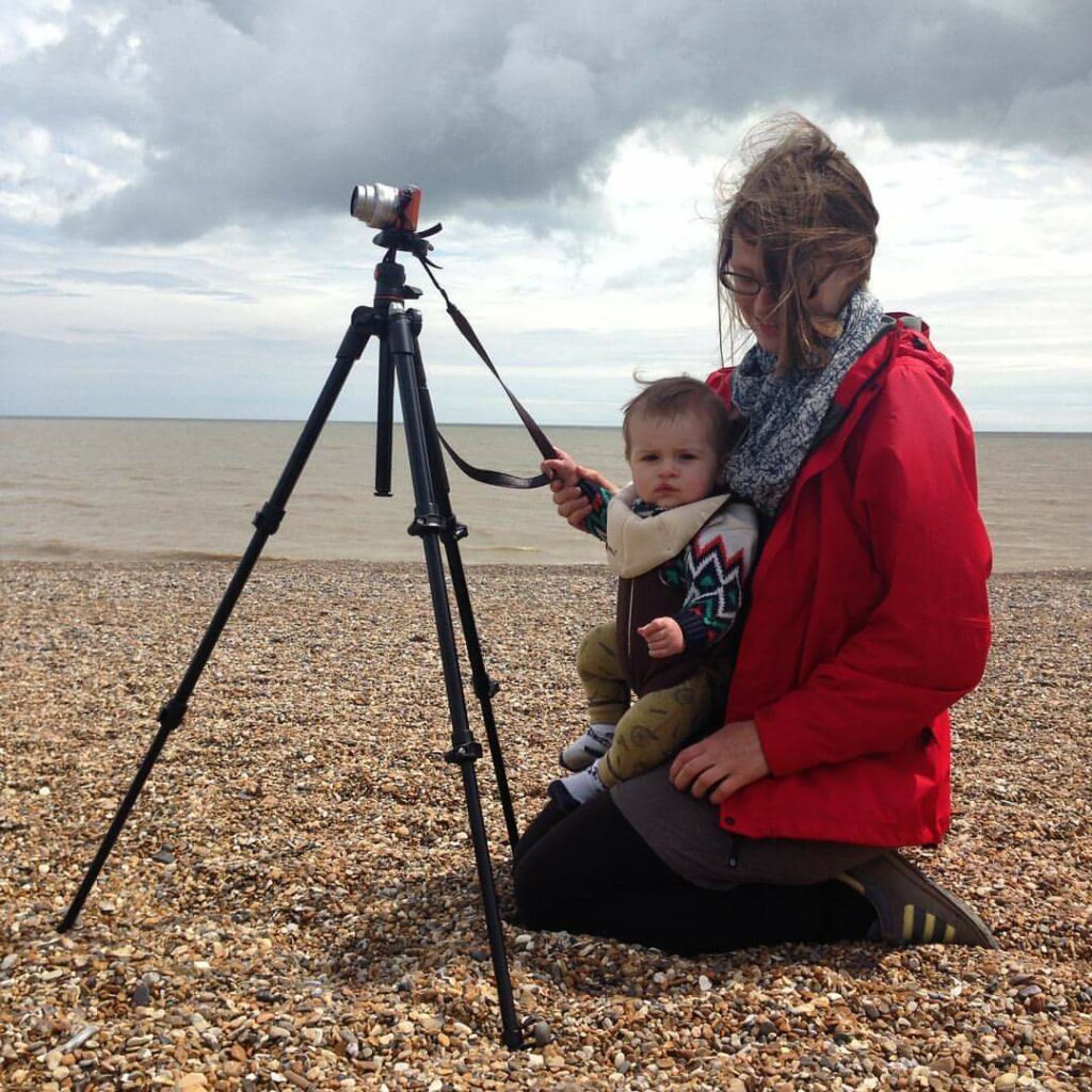 Photograph of a women in a red coat with a baby in a sling kneeing in front of a tripod with a camera on it. 