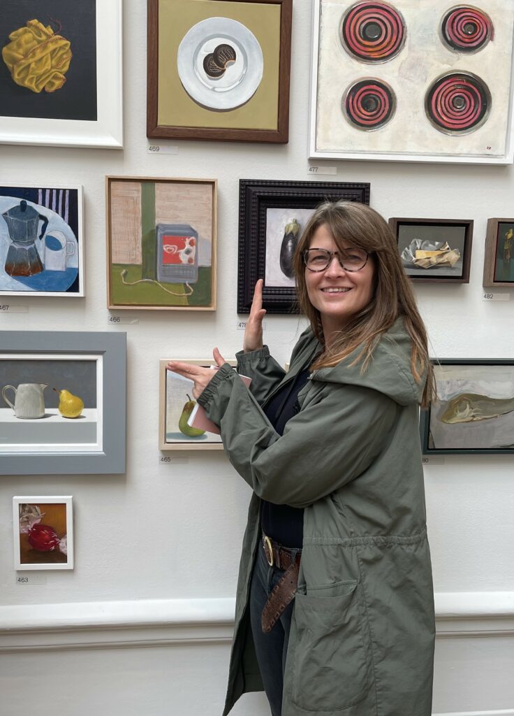 Photograph of a lady stood in front of a wall of paintings pointing at the painting she made
