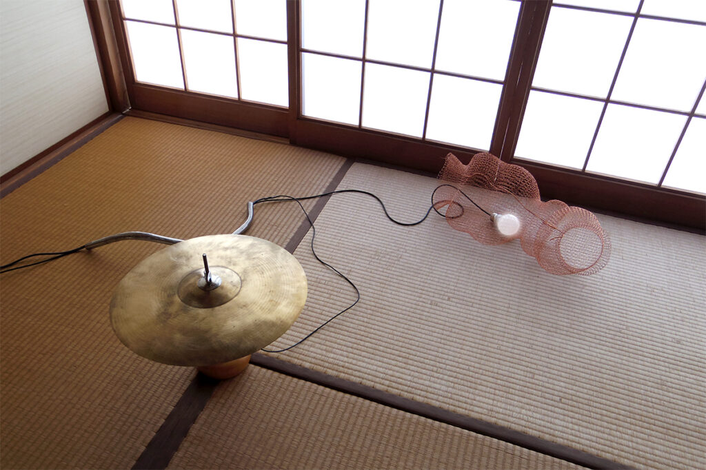 a photograph of an artwork installed on a carpeted floor. The artwork is made up of a symbol from a drum-set, a lightbulb that's turned on and is sitting inside a tubular copper mesh structure.