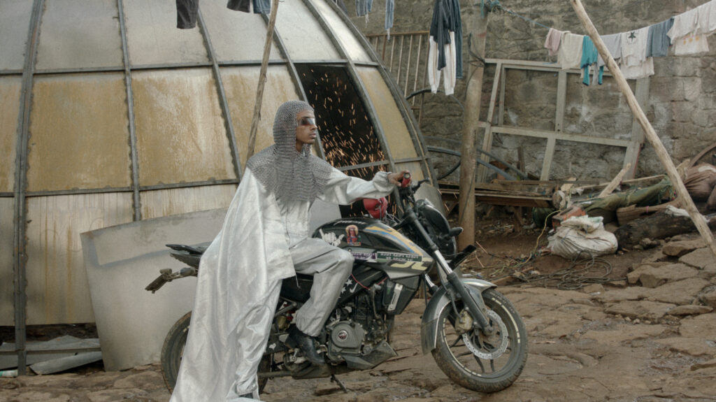 person in chainmail and cape sits on motorbike with sparks in the background