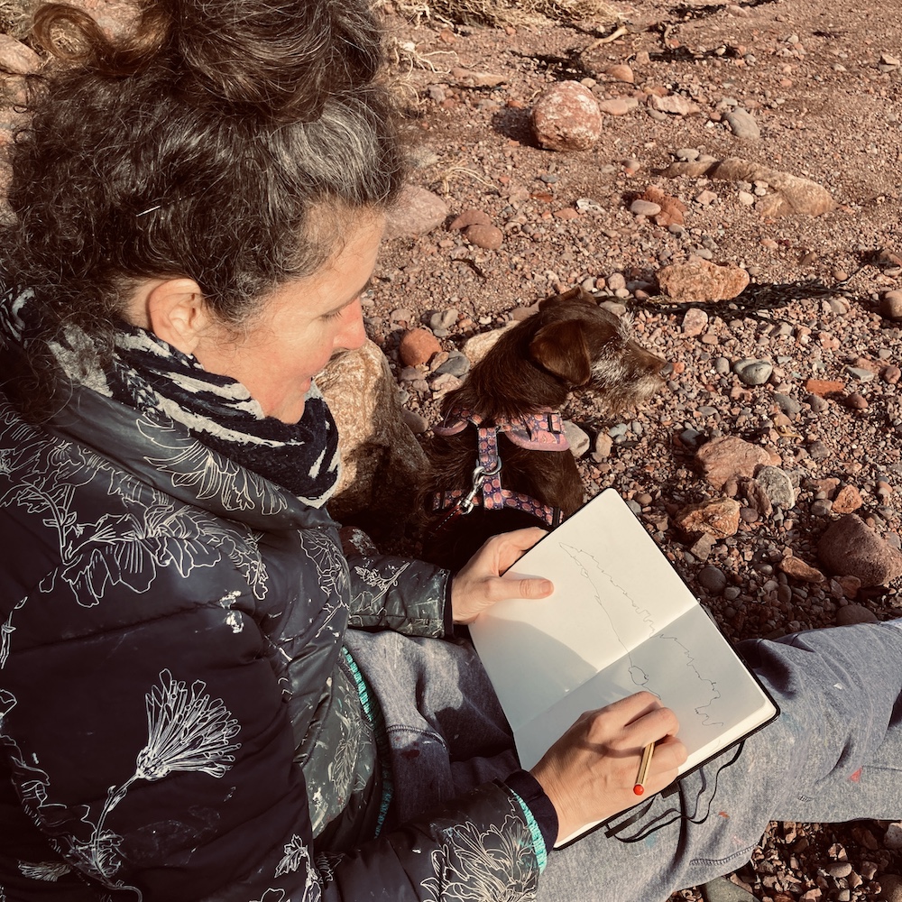 Photograph of a lady i na coat sat on rocky ground with a dog drawing in a sketchbook.