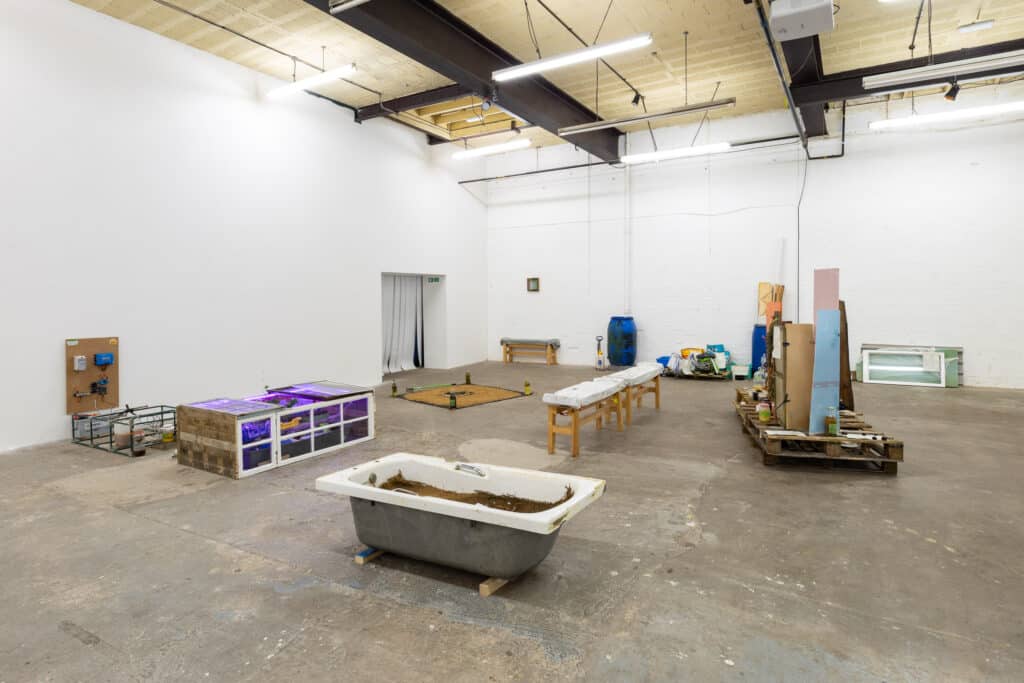 Photograph of an arrangement of art works made from reclaimed windows, a recycled bath and reused wood in an industrial building with white walls and concrete floor