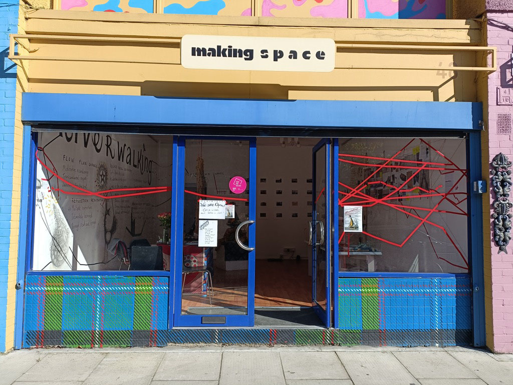 Photograph of a blue and yellow shop front with large windows and a sign titled "Making Space" above the front door. Looking through the open door and windows we can see a variety of mixed media artworks installed in the space, including one art work that is red tape taped across the windows.