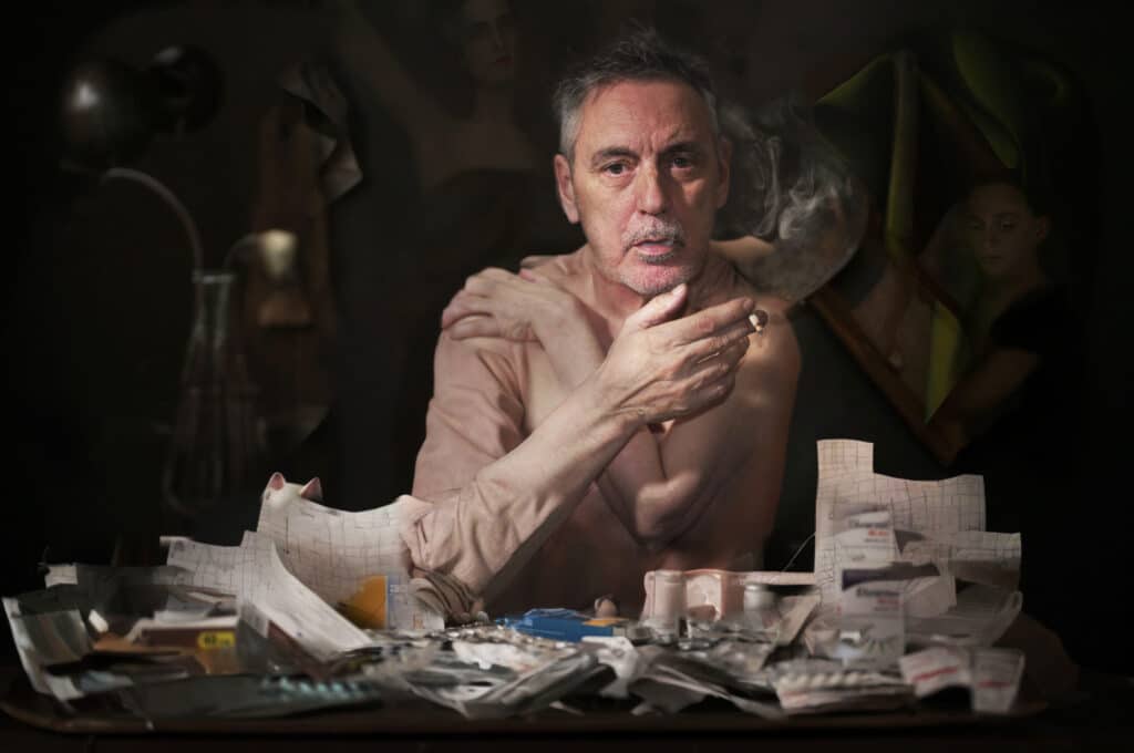 Photogrpahic portrait of a man sat at a table smoking, surrounded by paper and medication packets