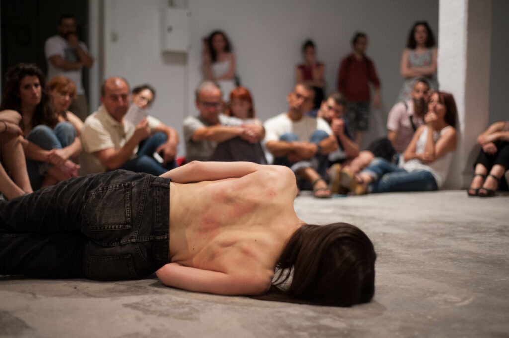 a photograph taken behind a half naked person who led on the floor in front of an audience that sits on the floor