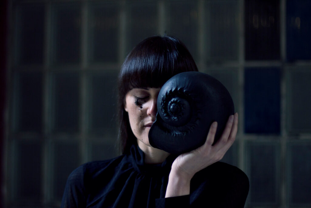 portrait of a lady dressed in black with black hair holding up a large black shell that covers half of her face