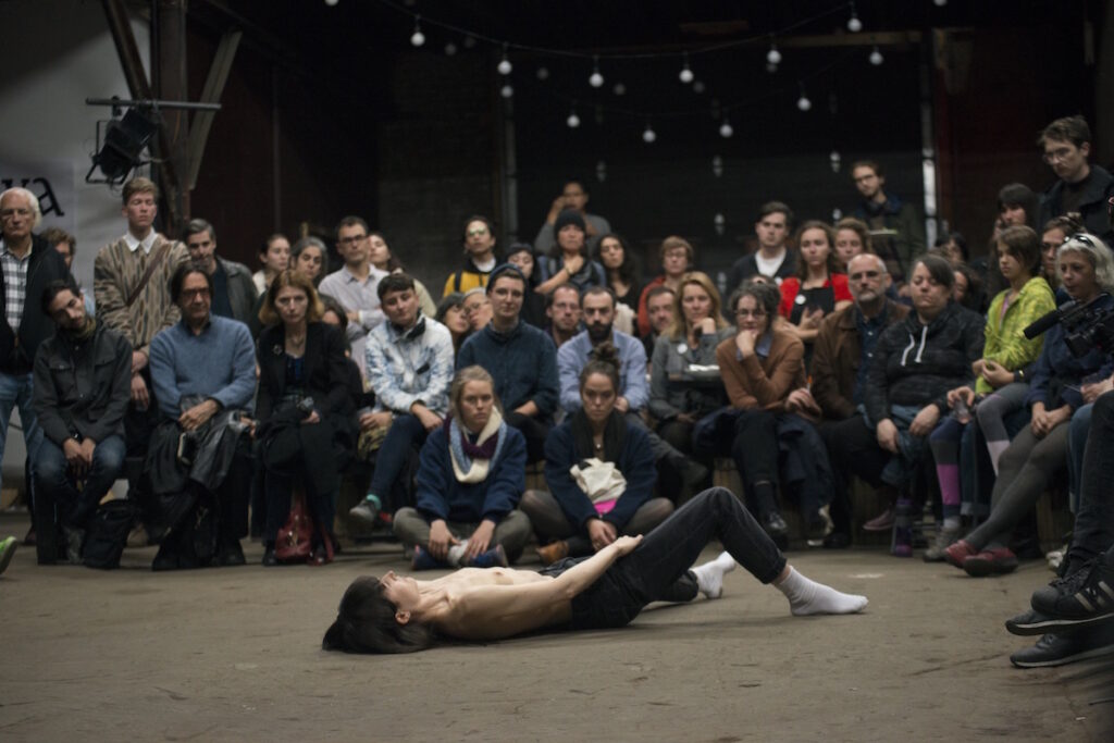 Photograph of a half naked person led on their back on the floor in front of a sitting and standing audience
