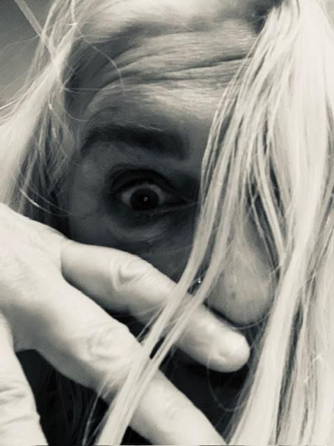 [black and white photo of a face with long white hair, hand over mouth and only one eye visible]