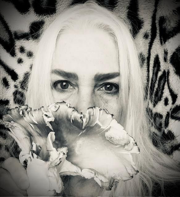 [black and white photo of a face with long white hair and an oyster mushroom held in front of the mouth , against a leopard print fur background]