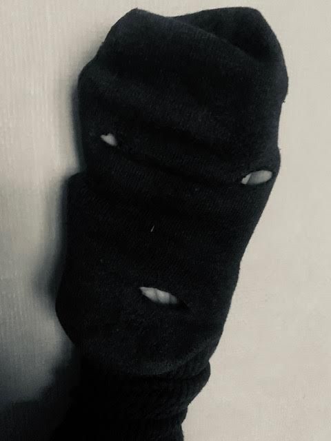 black and white photo of a dark sock stretched over a hand so it looks like a face, with three holes that suggest two eyes and a mouth, the heel is a chin and the toe could be ears