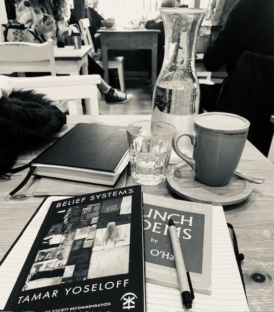 black and white photo of a cafe interior with a collection of things on a table top: an empty coffee cup, a bottle of water, a half full glass, a notebook and pen, a couple of sketchbooks, a diary, two books of poetry: ‘Belief Systems’ by Tamar Yoseloff and ‘Lunch Poems’ by Frank O’Hara