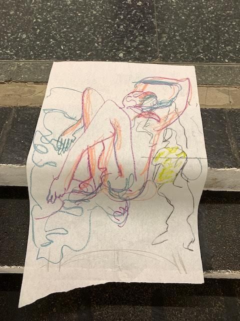 Colour photo of a life drawing of a reclining figure with hand above head. The drawing is on a large sheet of off white paper which is folded over two grey stone steps
