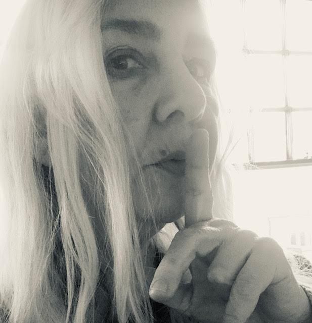 Black and white high contrast photo of a head with long pale hair, face in half profile, finger held to mouth, strong light and window pane to right hand side of image.