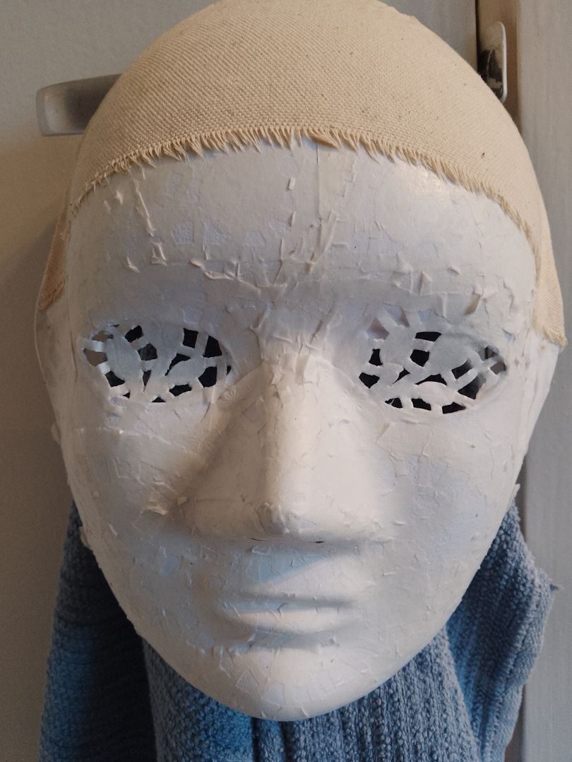 A paper mask covered in white paper doily lace with a cream canvas cap.