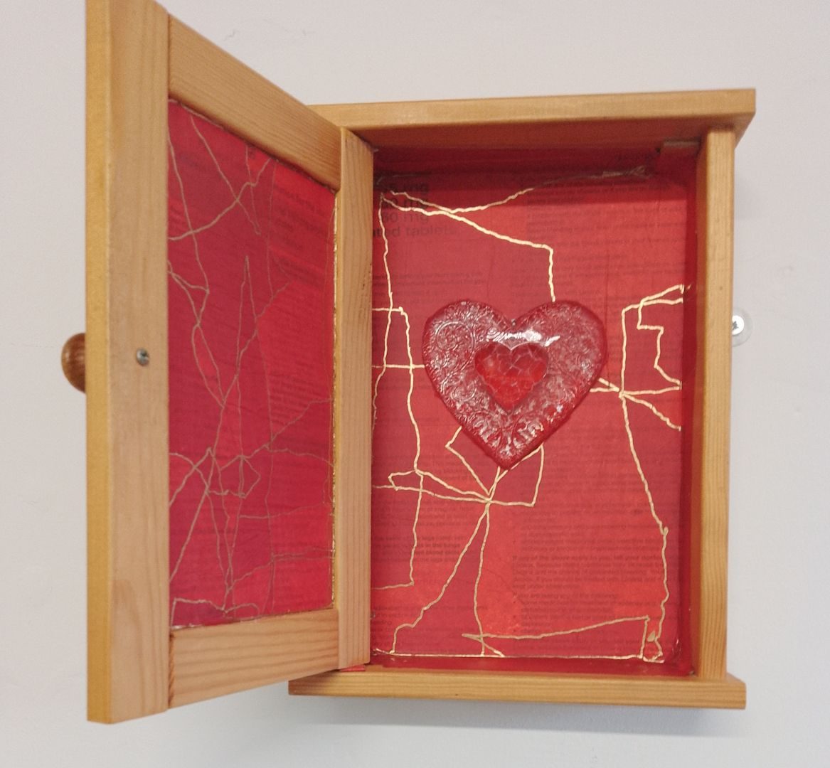 Asmall wooden cabinet with its door open. The inside is lined with red collage with gold ink lines around the edges of the tron material. A heart-shaped Christmas clear plastic ornament hangs in the centre.