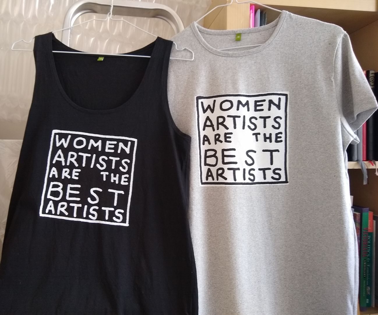 A photo of a black vest and a grey T-shirt on white wire cothangers. Both t-shirts have the handwritten slogan 'WOMEN ARTISTS ARE THE BEST ARTISTS' printed in a square on the front. The black vest has white text with a white border, the grey t-shirt has black text and a black border on a square white background.