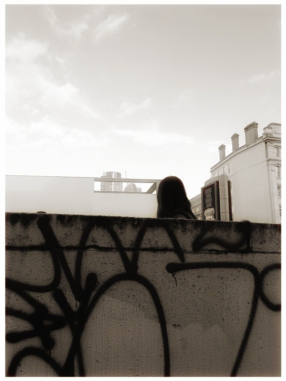 A sepia tinted photograph. A person's head seen from the incline of an underpass. The picture is broken up into bands - a graffitied wall, the person's head and distant buildings and the sky.