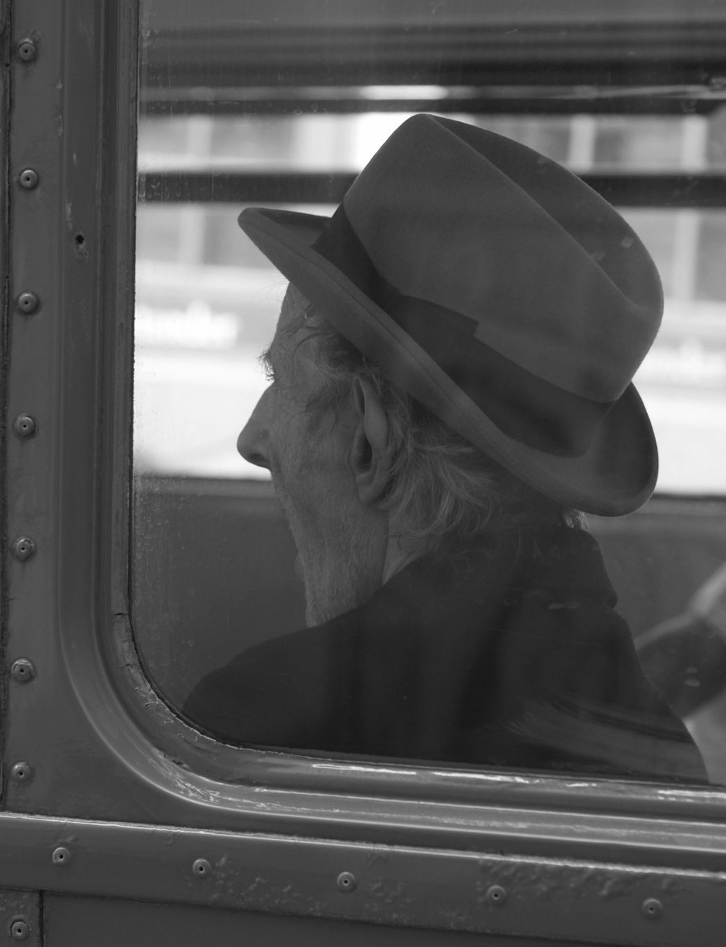 Black and white photograph of a man's (hatted) head framed in the window of a bus