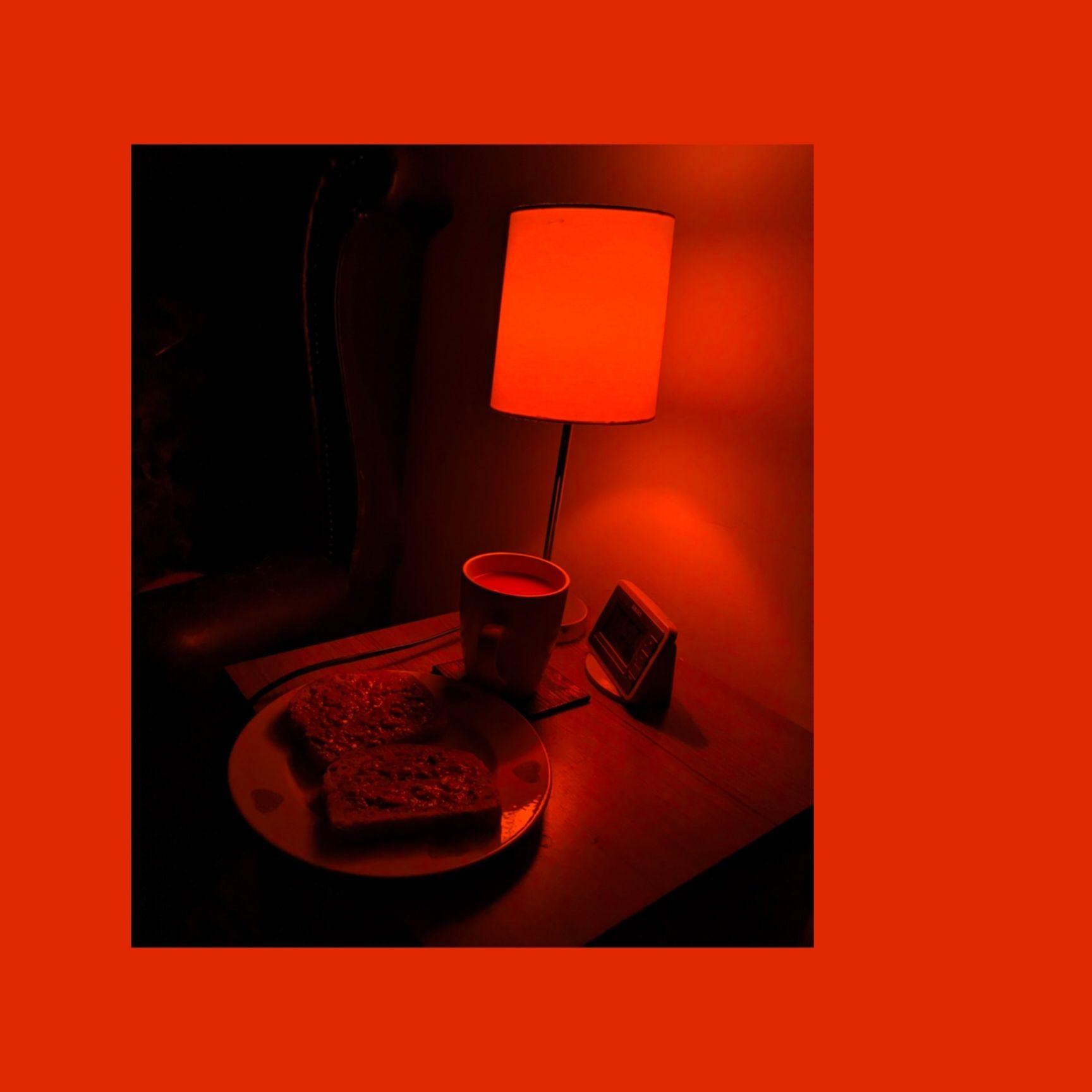 A dimly lit still life photograph of a bedside table with a  cup of tea and two slices of toast under an orange desk lamp.