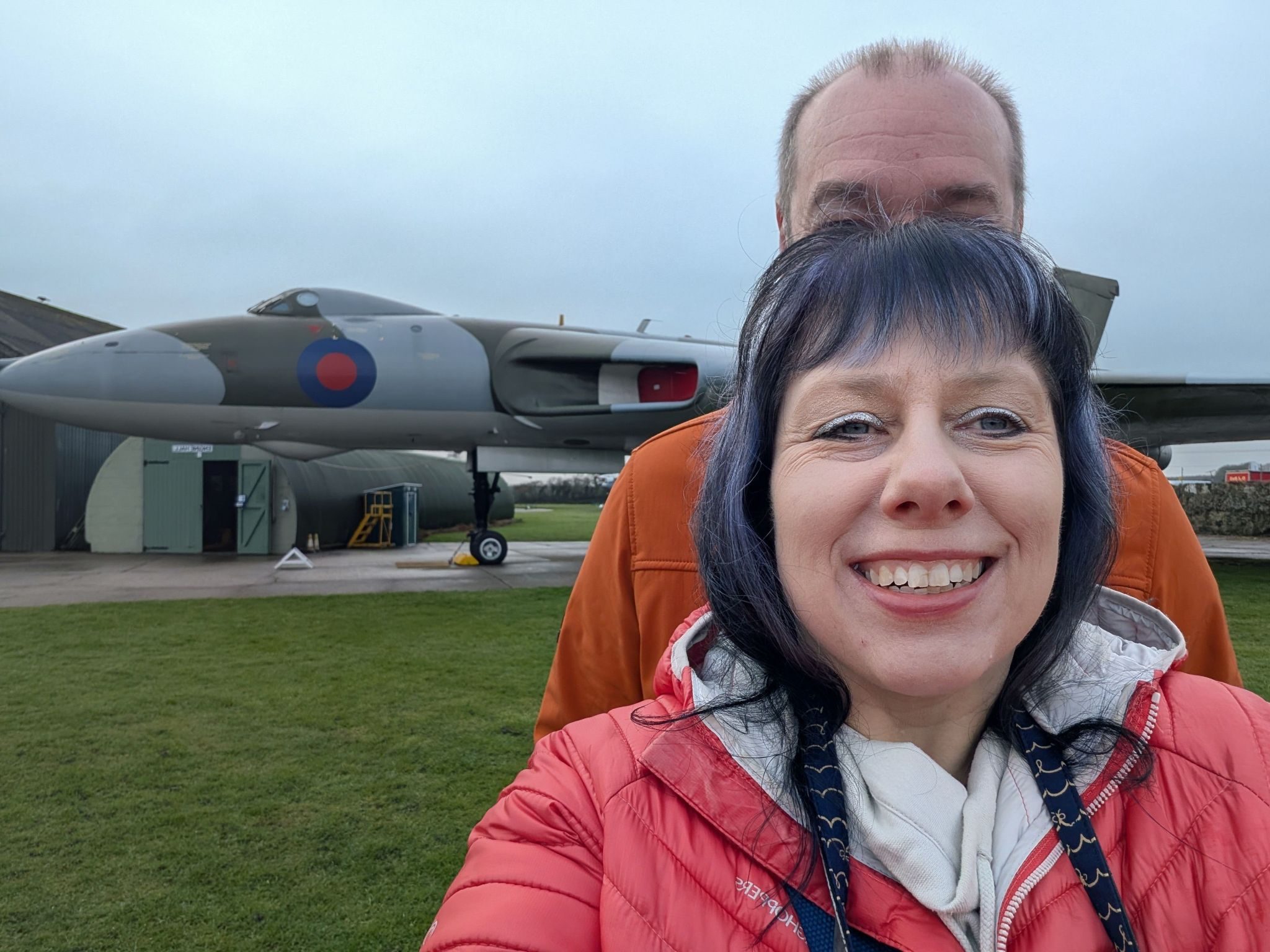 Day out with husband hiding behind me at an aviation museum with the avro Vulcan in the background. Clear sky and grass. I have black and purple hair and am wearing a coral winter coat.