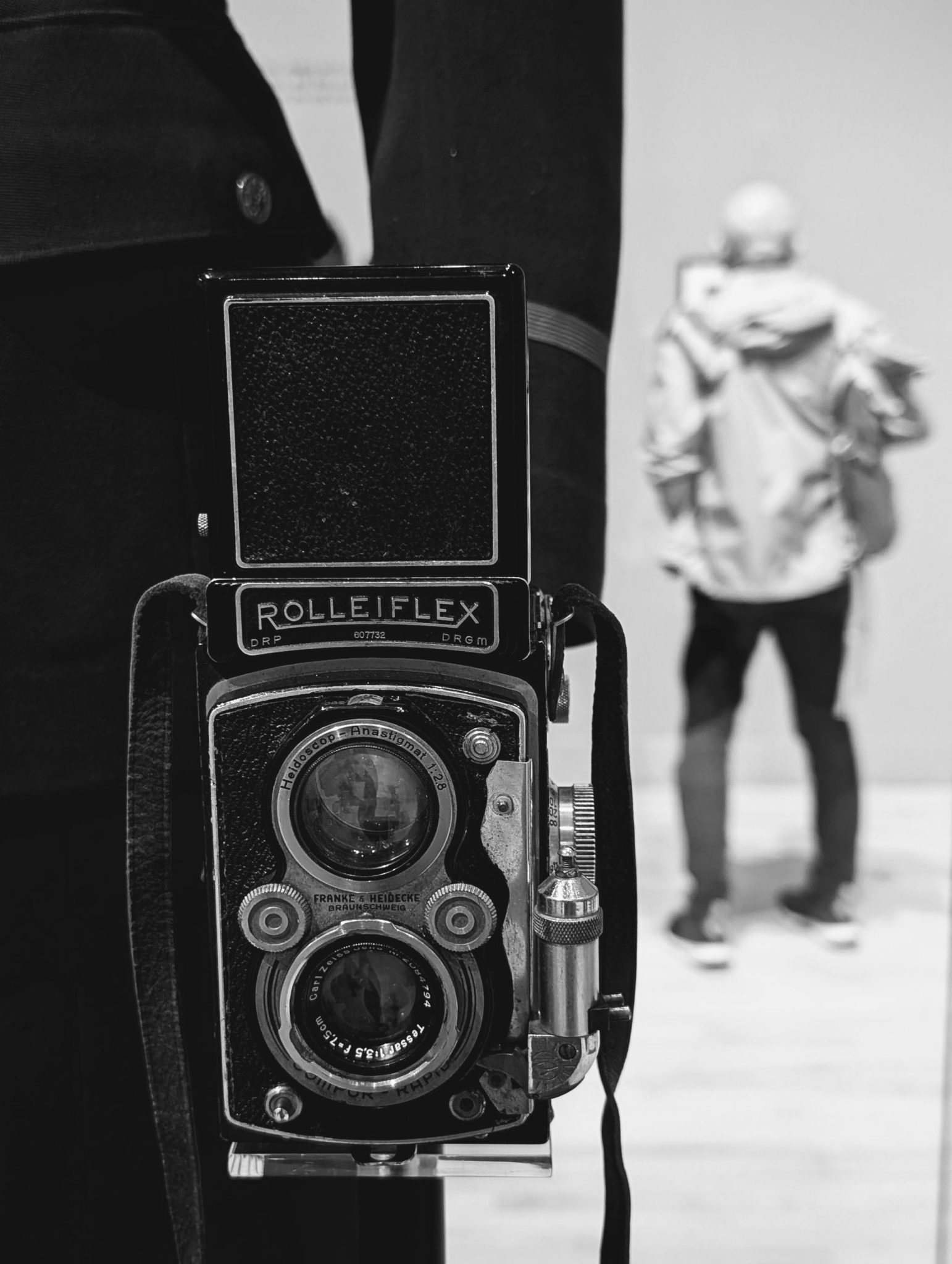 Black and white photo of a display cabinet with a Rolleiflex camera and a man on the background looking at the works