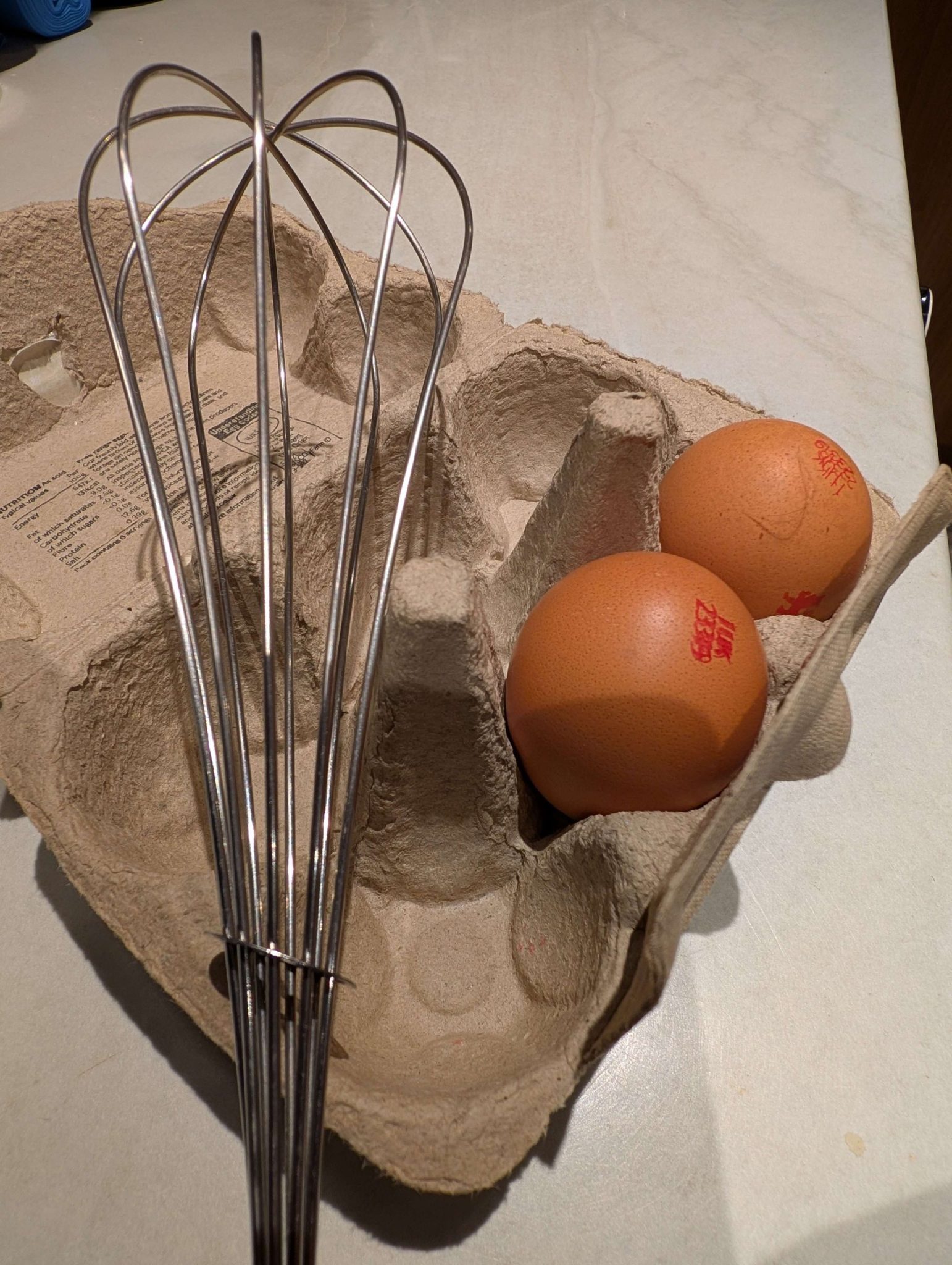 Two eggs in an egg box and a whisk