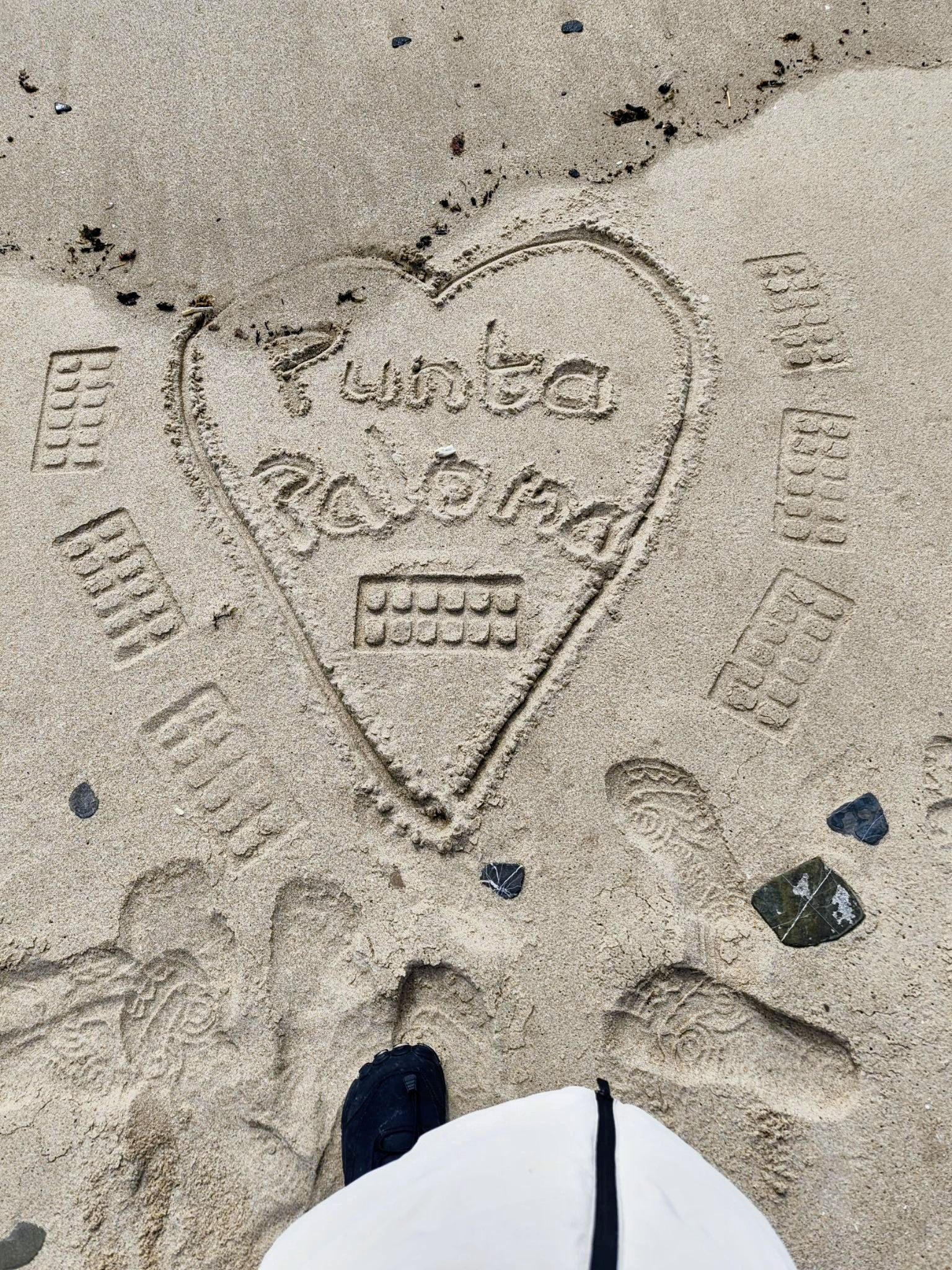 In the sand a drawing of a heart shape with the words Punta Paloma written inside, and patterns and the artists foot print and foot