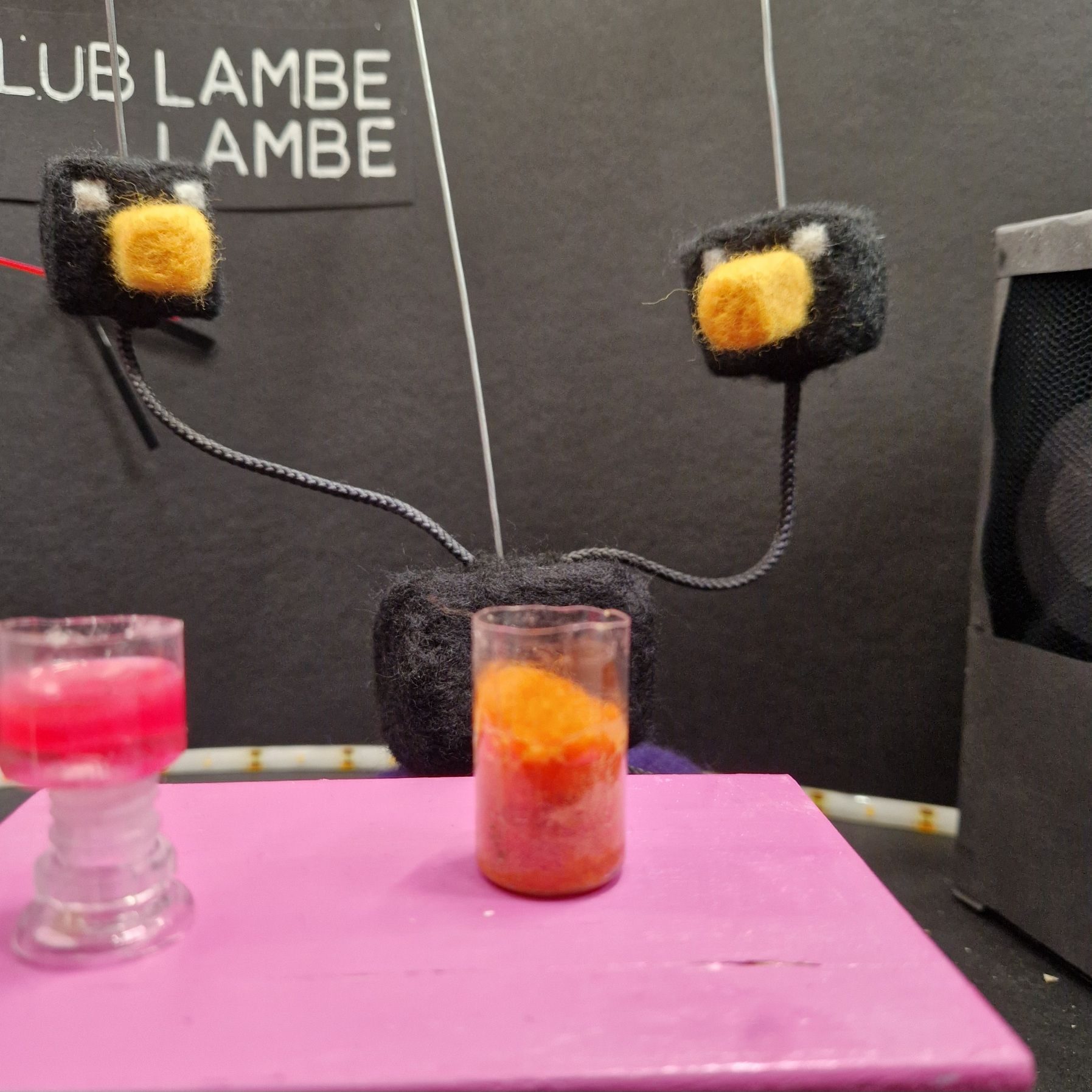 Two-headed black bird with yellow beaks dances. In front of it is a pink table with two drinks on it.