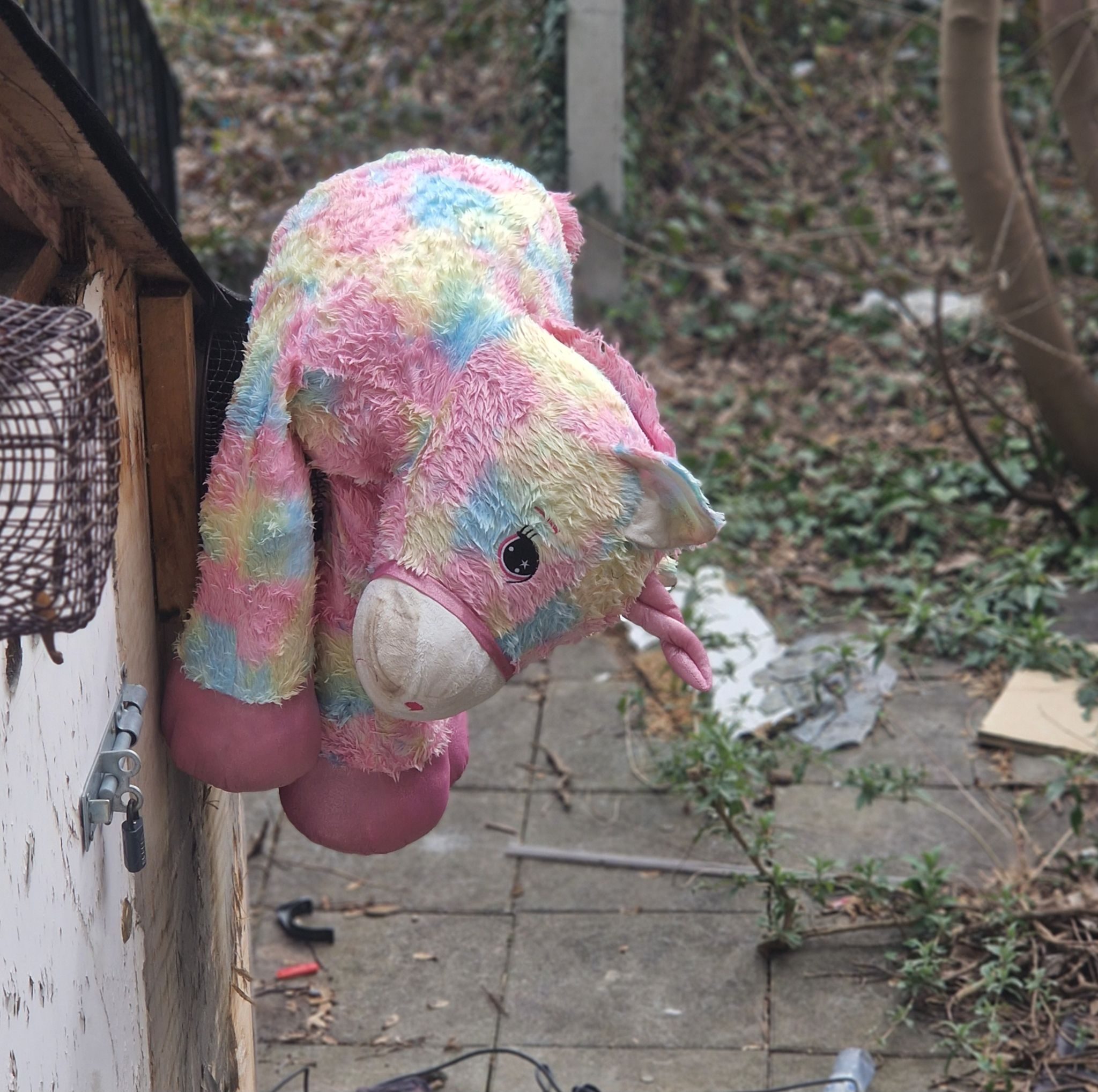 Pastel blue, yellow and pink unicorn soft toy slumped awarkwardly by the roof of an outbuilding