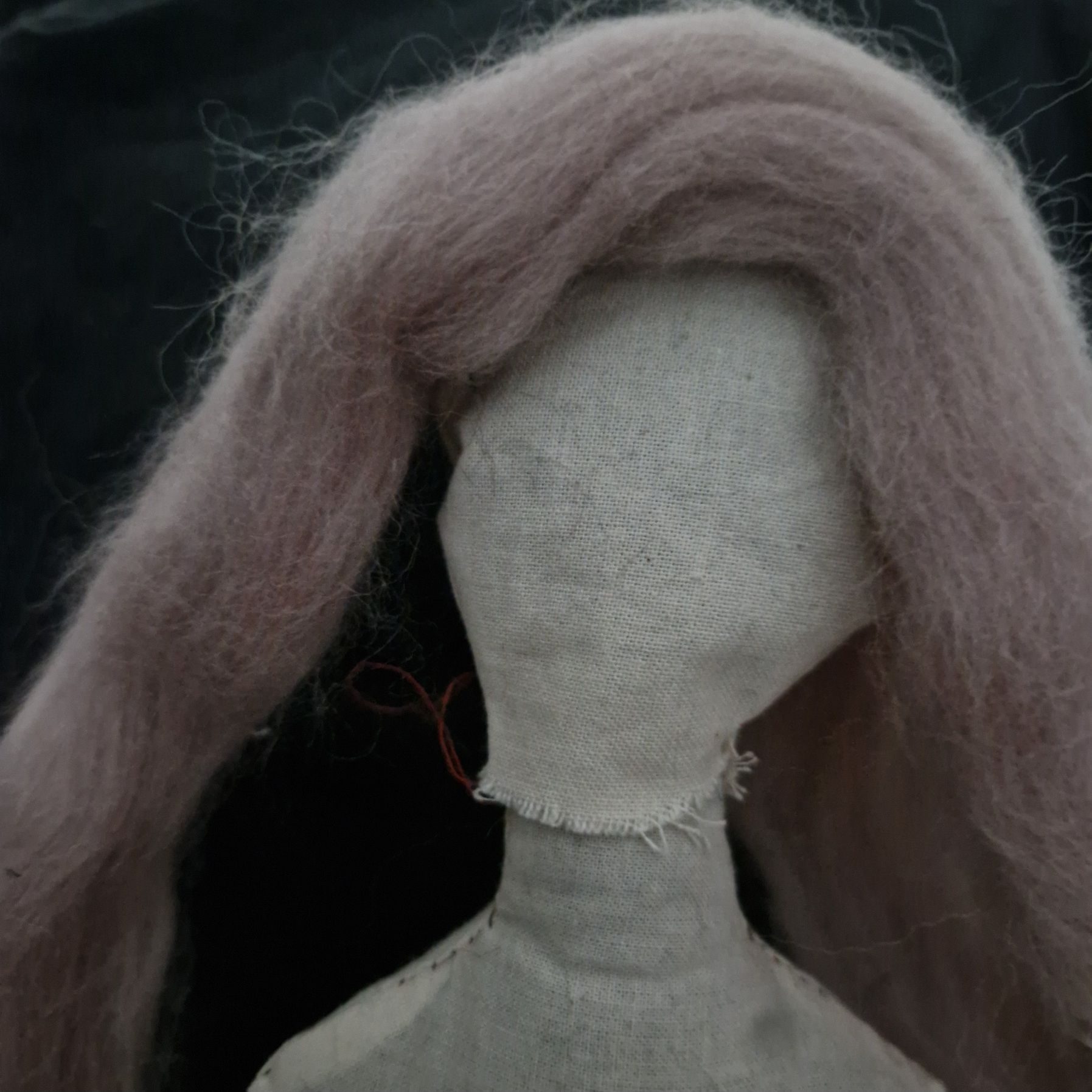Portrait view of natural coloured cloth doll. The doll has no facial features and it has long shank of silvery pink wool plonked on its head in place of hair