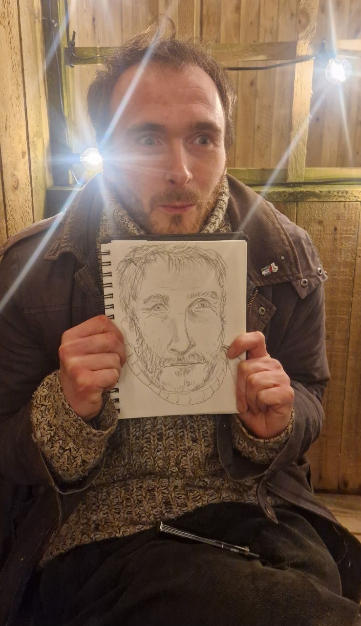 A man pulling a face holding a pencil self portrait in a spiral bound book