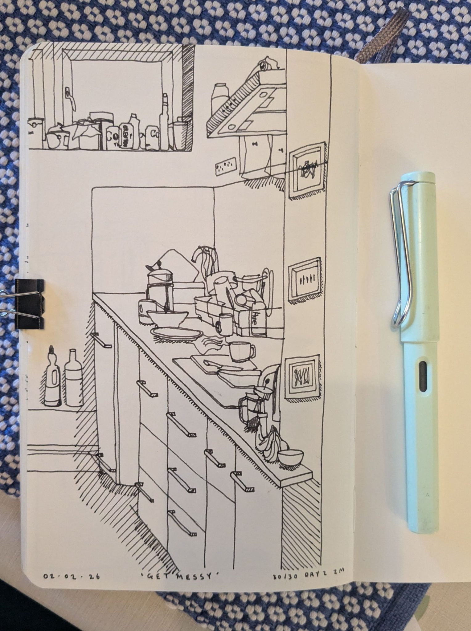 Photograph of a sketchbook page on a blue and white background. The sketchbook shows a pen and ink sketch of a kitchen. There is a pen next to the page.