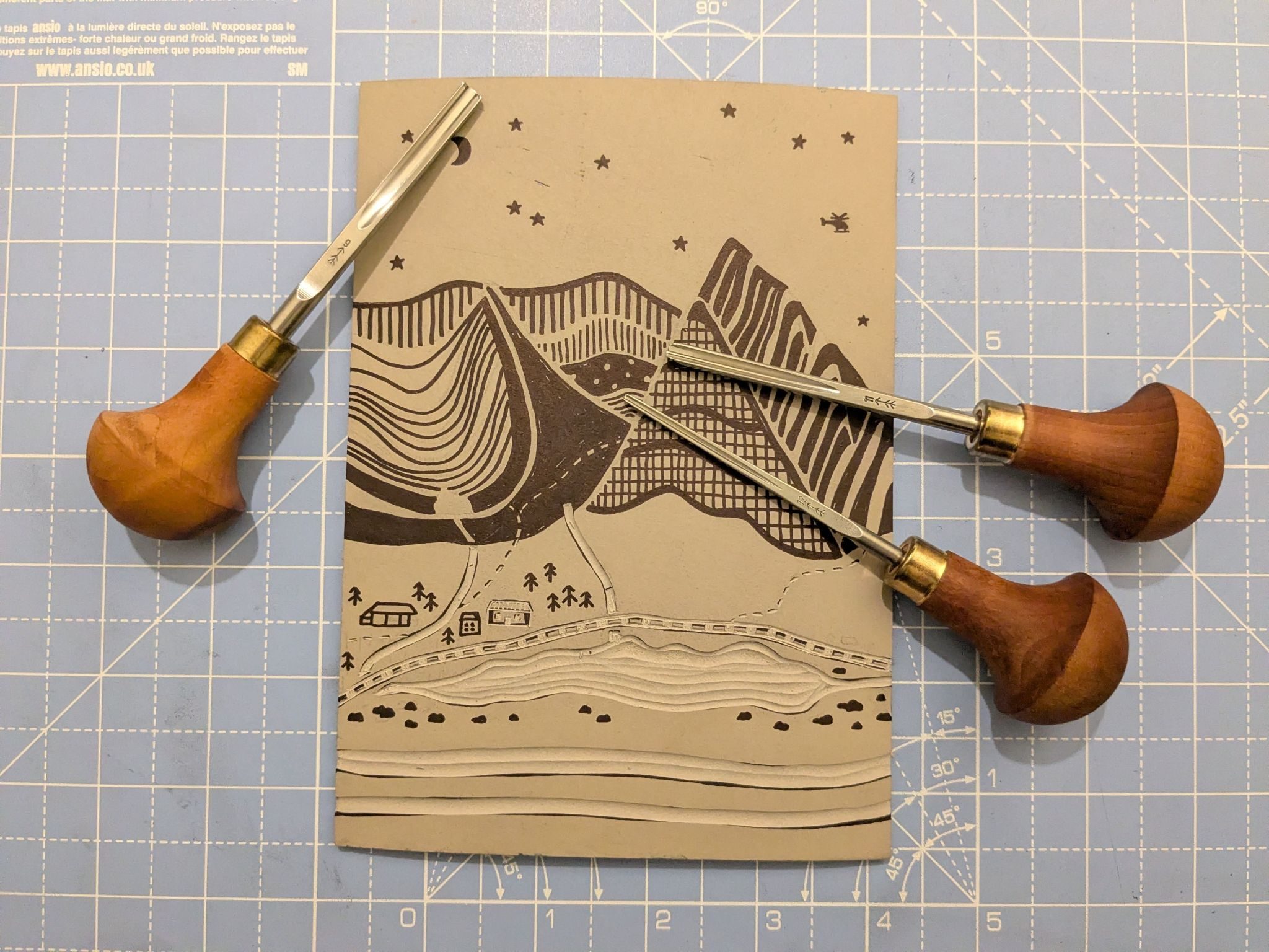 Photograph of a half finished lino carved block and tools on a blue cutting mat