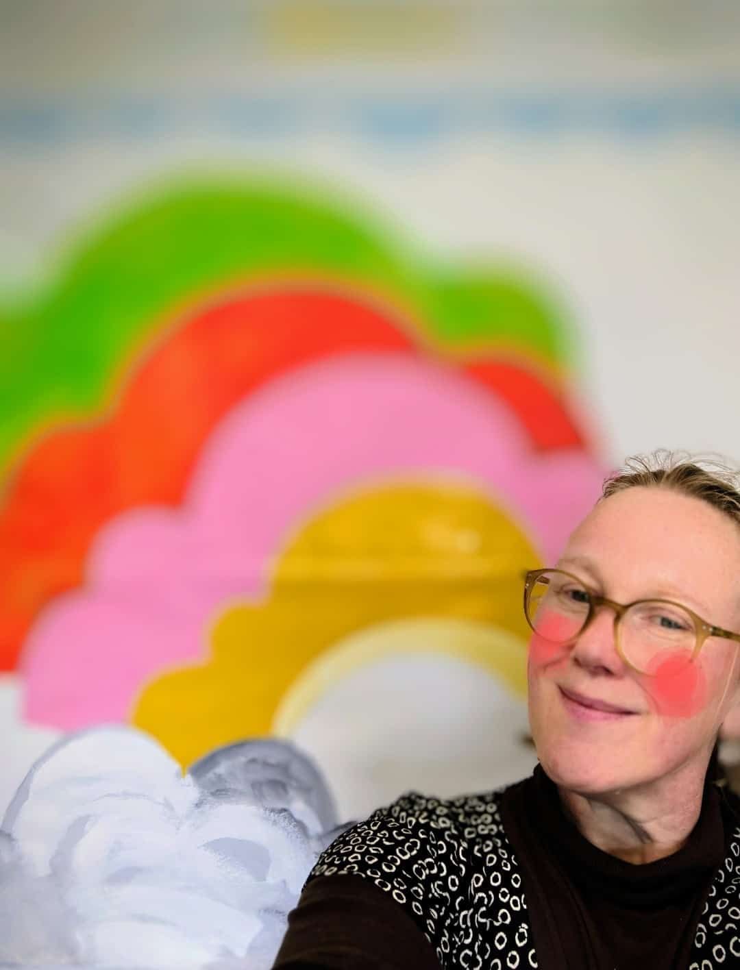 Woman, with rosy cheeks in front of a painted rainbow