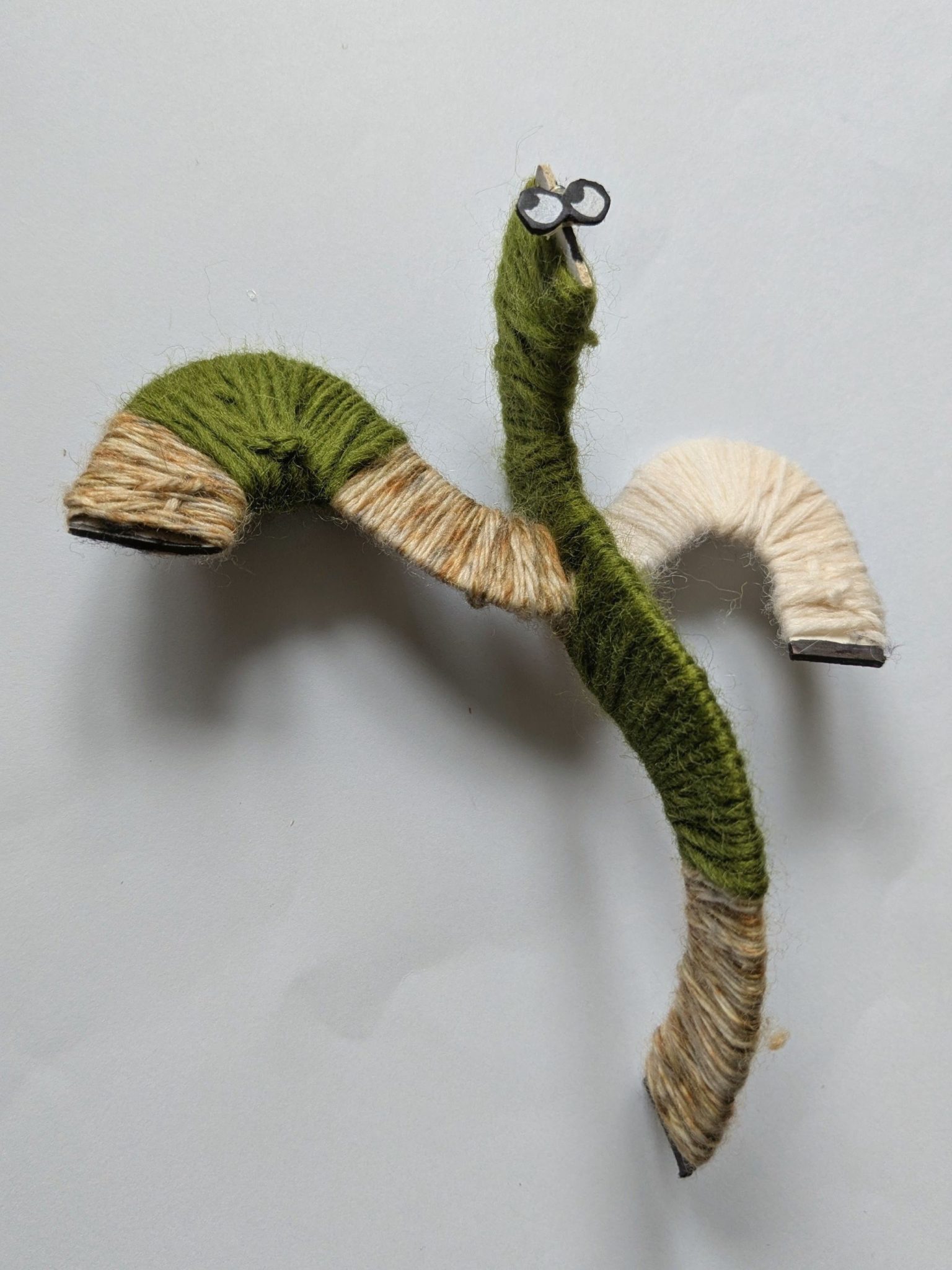 An abstract bendy figure with two eyes looking to the left corner. Woollen body made up of green, brown and white wool.