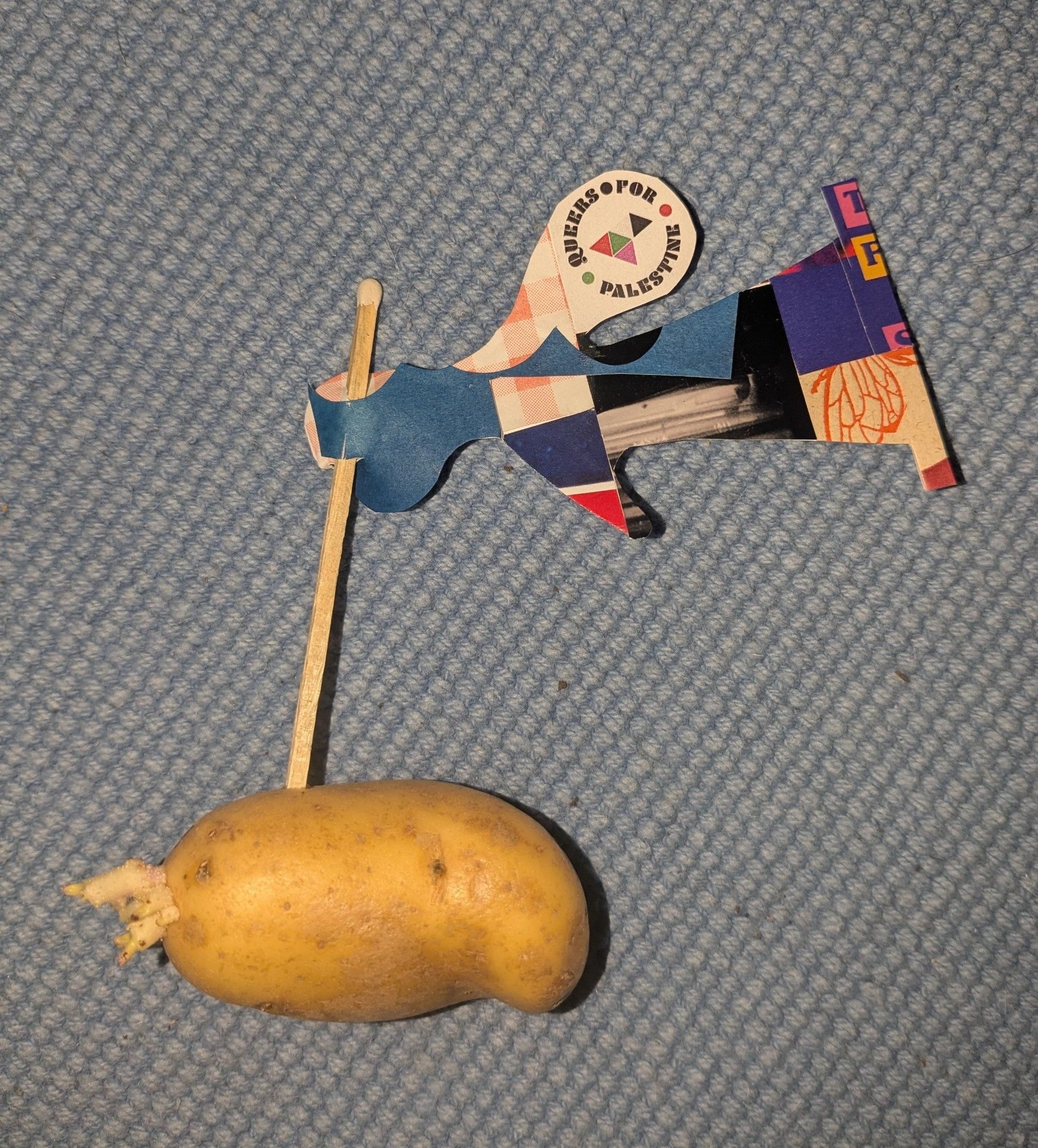 Flag made from collaged paper and Queers for Palestine leaflet. Flag post made from a giant matchstick and stuck into a small potato.