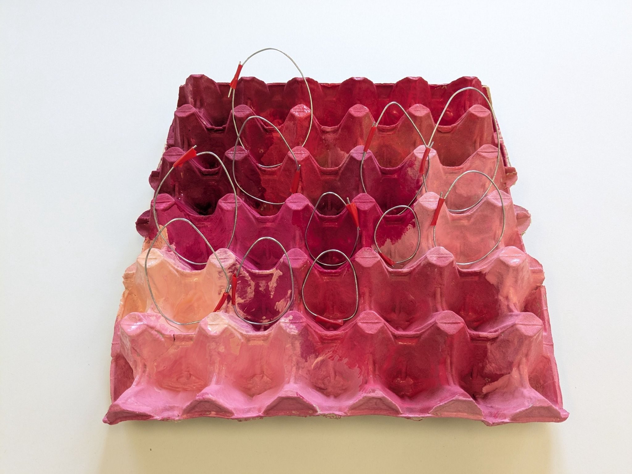 A pink painted large egg tray featuring different shade of pink. Shapes of eggs made from metal wire sit in the egg tray. The tray is on a white background.