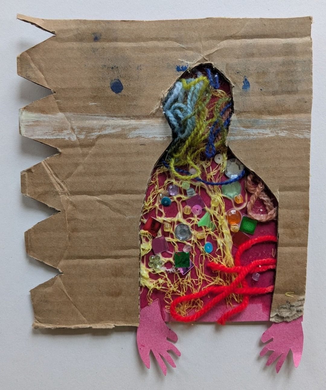 The shape of a torso and head is cut out from a piece of cardboard and filled with different coloured wool and sequins. Two pink hands fall at the bottom sides of the figure.