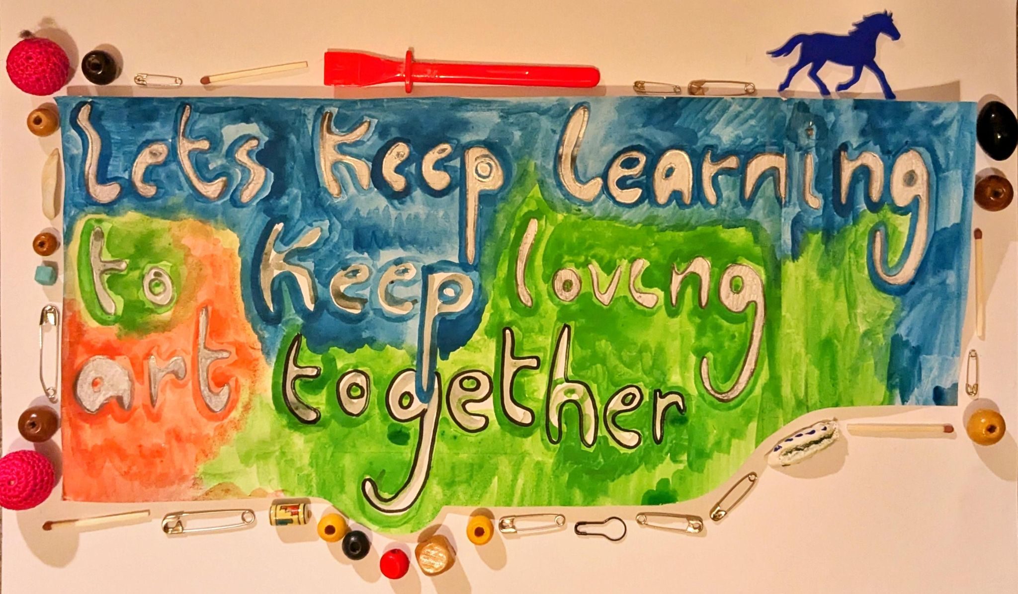 A painted piece of text using blue, green and red watercolour reading 'lets keep learning to keep loving art together'. The piece of paper is circulated by a border of safety pins, beads, matches and other random objects.