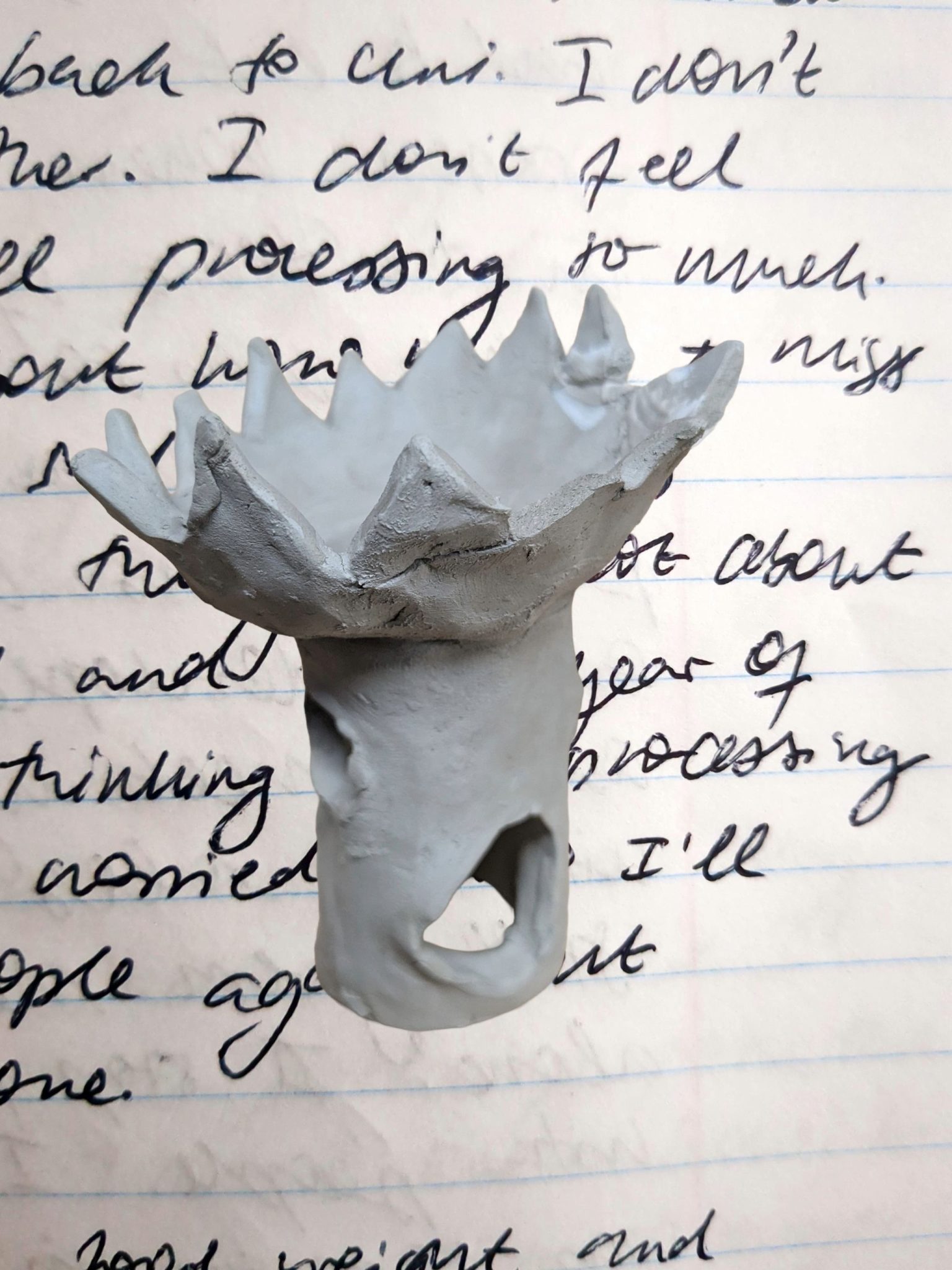 A clay object made from a tube with an open flower cup shape on top has been cut out and sits against a background of zoomed in handwriting on lined paper.
