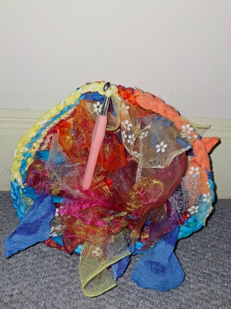 A basket-shaped crochet piece in progress made of numerous colourful pieces of scrap fabric. Many loose ends protrude
