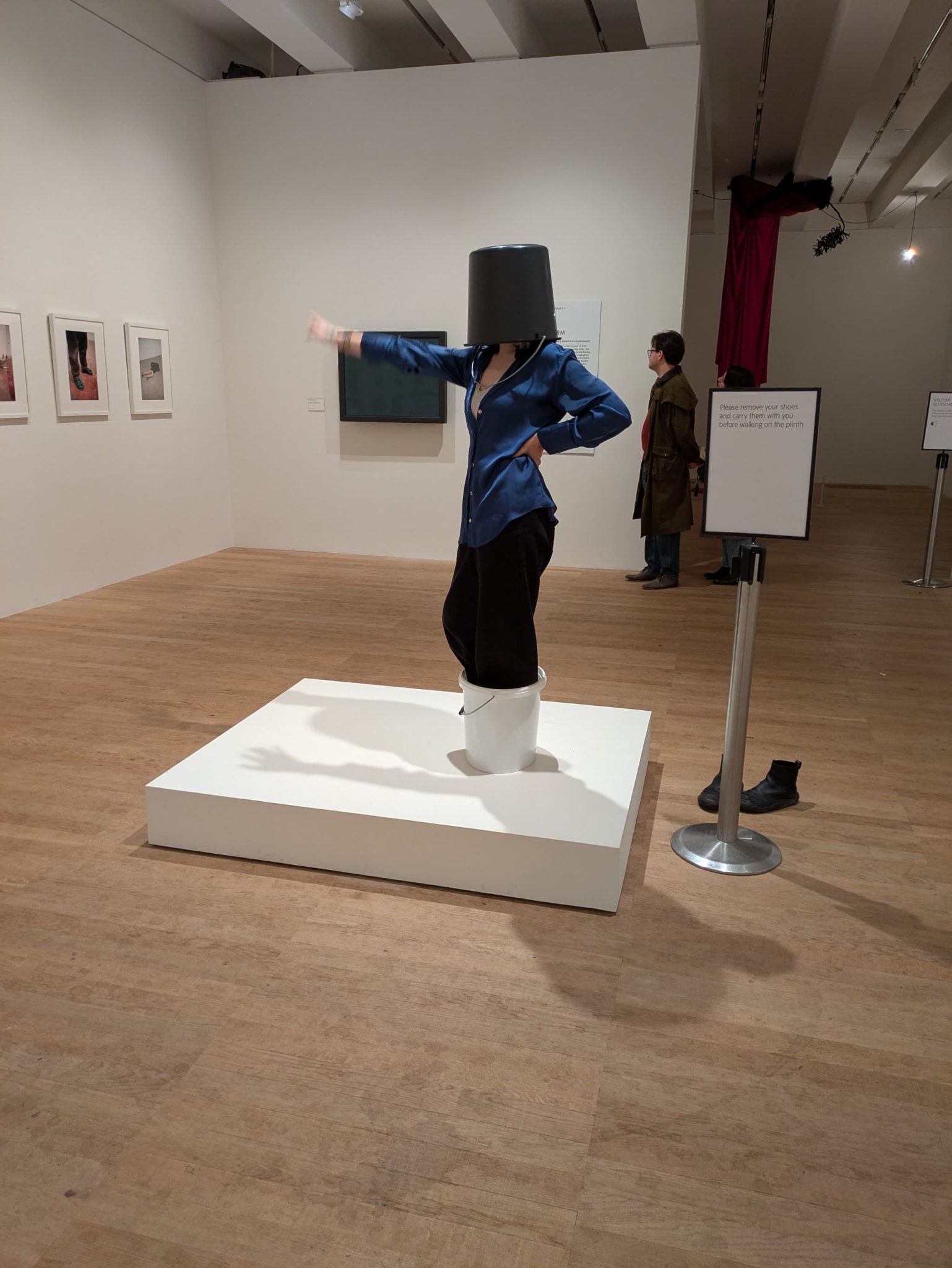 A woman in a blue silk shirt wears a bucket on her head and stands on a plinth inside a bucket