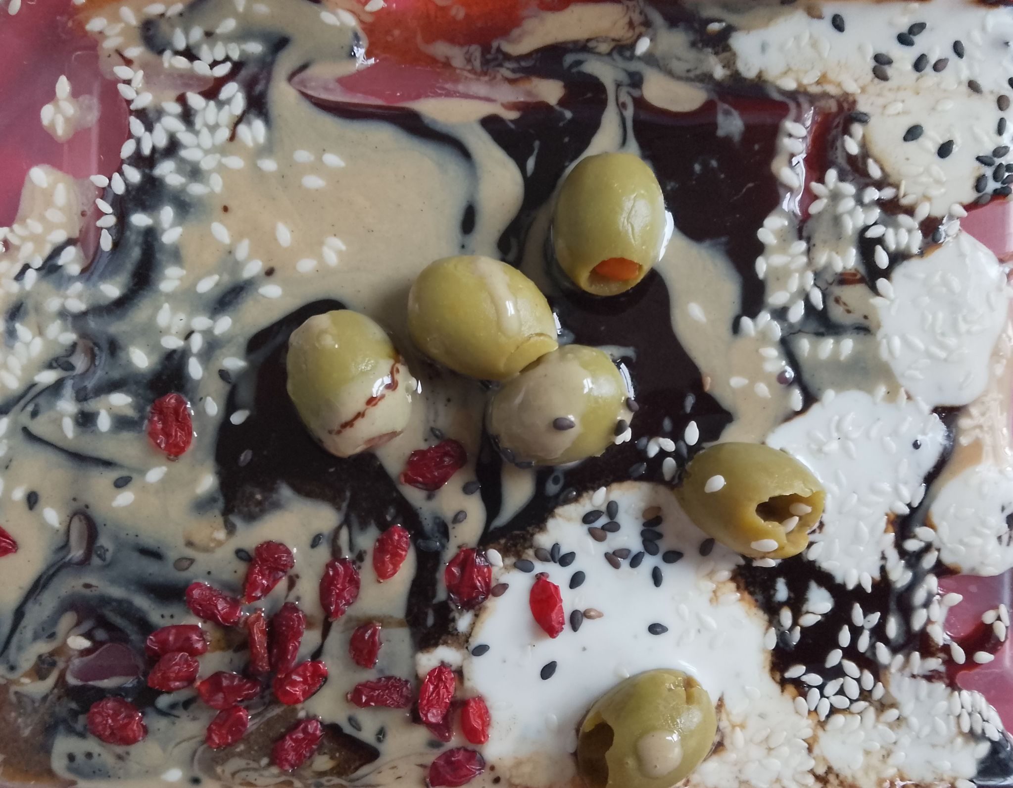 Abstract made with pomegranate molasses which is black, tahini which is cream yogurt sauce which is white with sprinkles of barberies which is red that green olives and sesame seeds