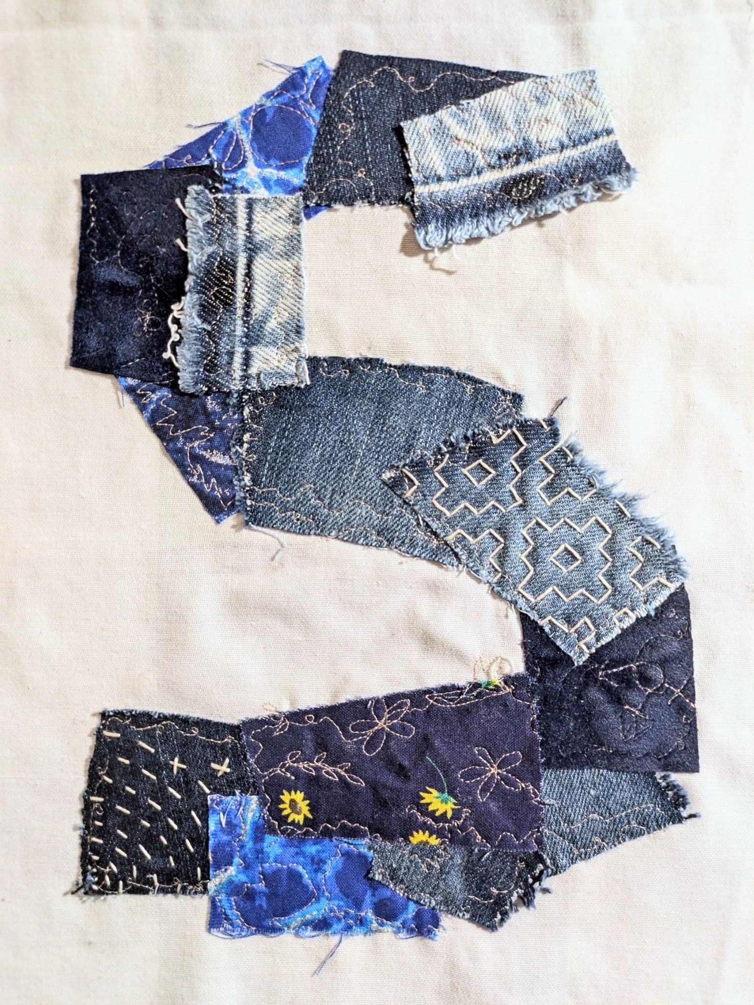 Blue and denim scraps of fabric stitched onto white ground in the shape of a large S, partly machine embroidery and partly sashiko