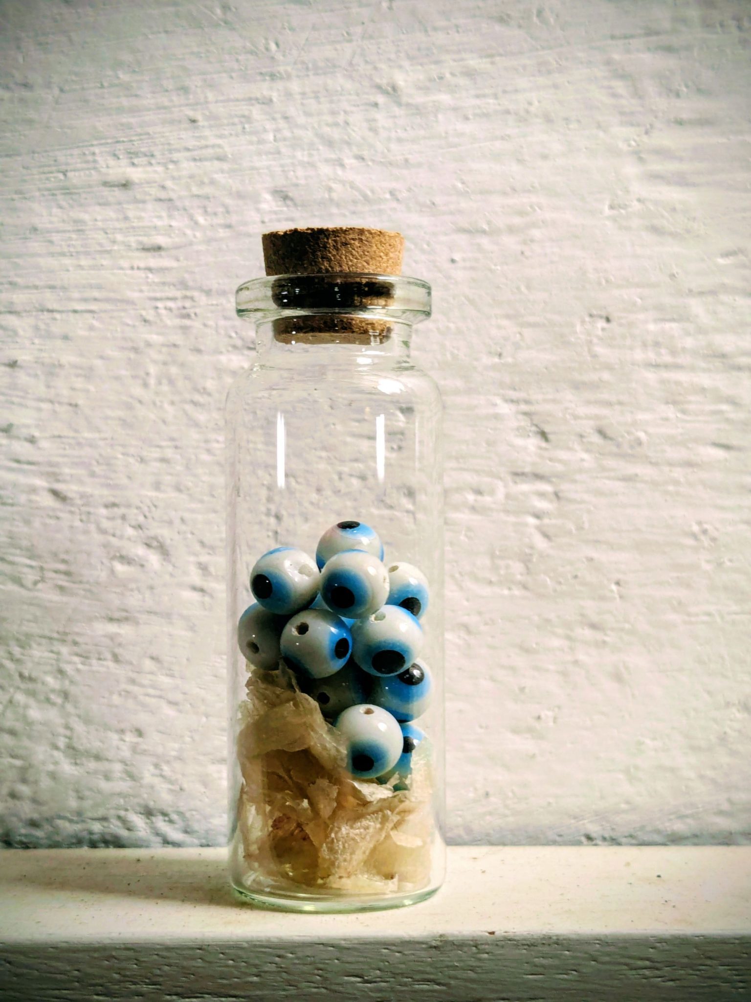 A small glass bottle with a cork stopper containing a layer of dry skin cells at the bottom, then some white and blue glass eye beads and the top third of the bottle is empty