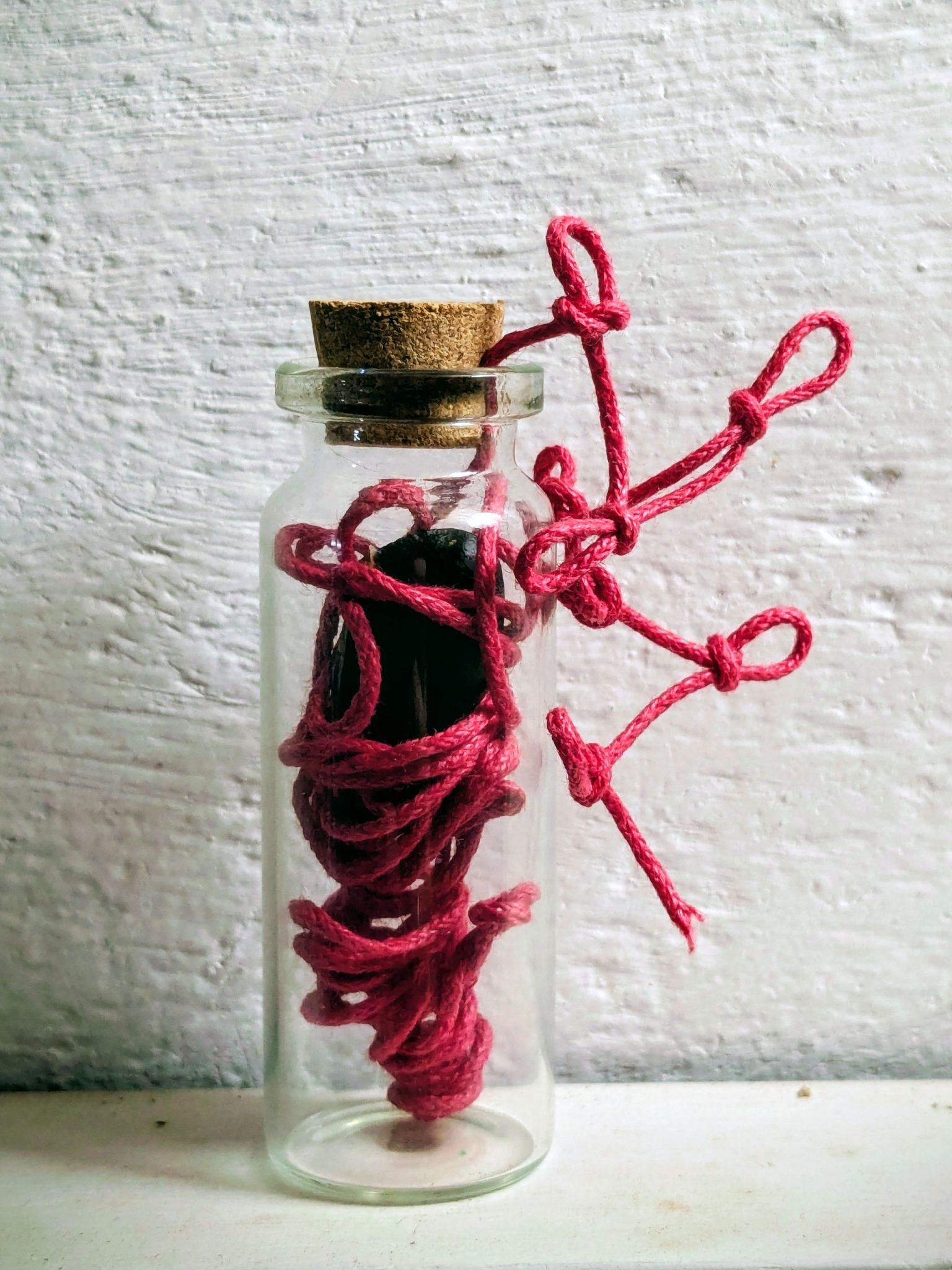A small glass bottle containing a chiku seed wrapped in red yarn, that comes out the cork stopper and evolved into 7 magic knots that looks like tiny leaves