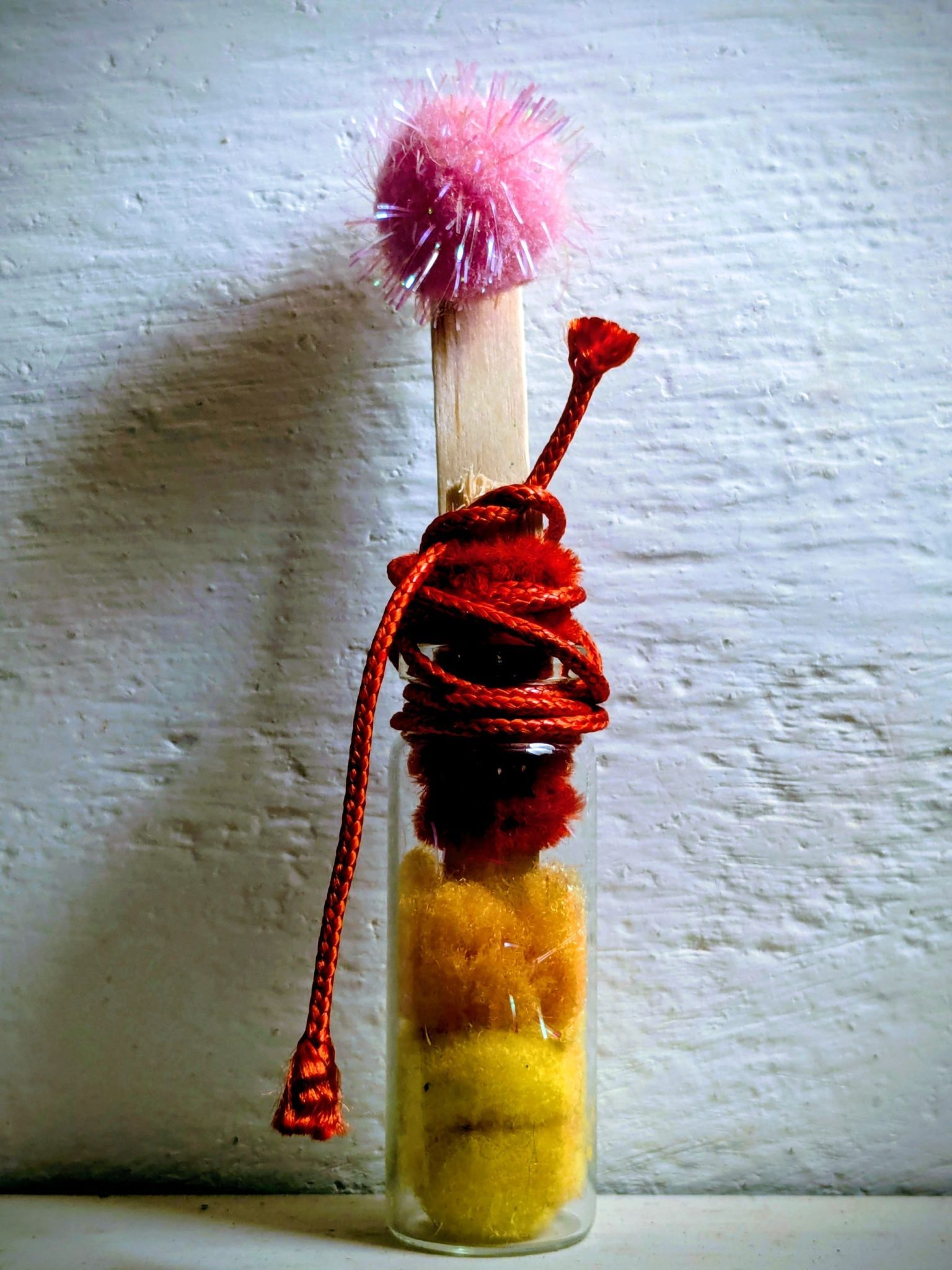 A small glass bottle containing orange and red pompoms, with the top wrapped in red yarn; an ice cream stick sticks out from the top & a pink pompom is glued at the top of the stick