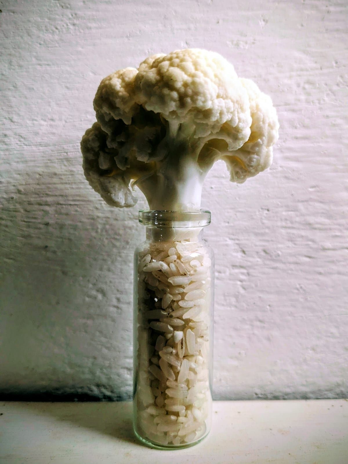 A small glass bottle filled with rice with a cauliflower floret sticking out of the top, looking cloud like