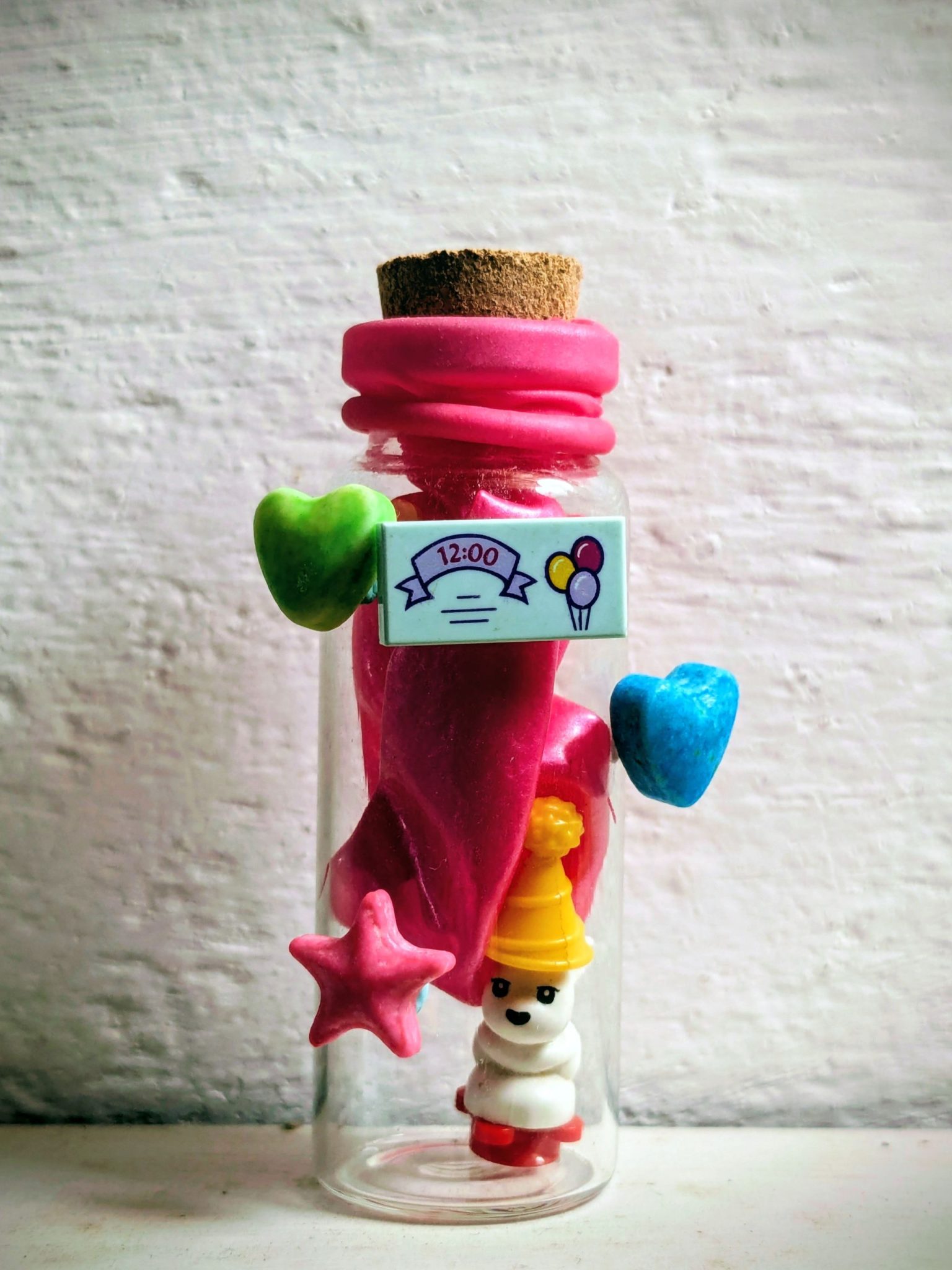A small glass bottle with a small plastic character and a pink balloon inside, with some colour candies bluetacked on the outside of the bottle and a small plastic sign with a drawing of balloons