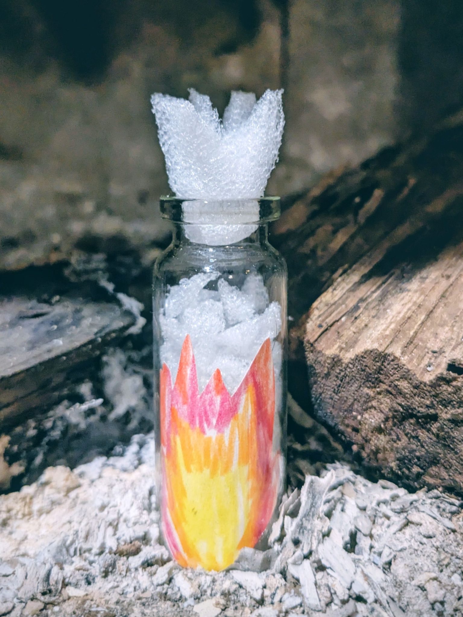 A small glass bottle containing a pencil drawing of fire and filled with pieces of styrofoam that look like snow; it has a styrofoam top shaped like a flame and the bottle sits in a background is ash & logs from a fireplace